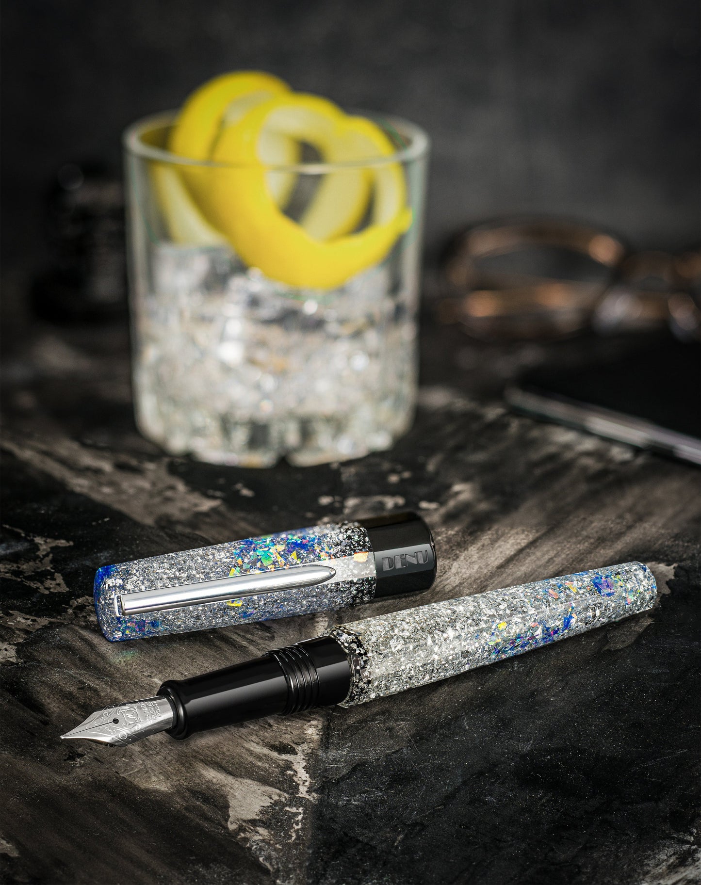 Fountain pen with decorative cap on a dark surface next to a glass of vodka and lemon twists